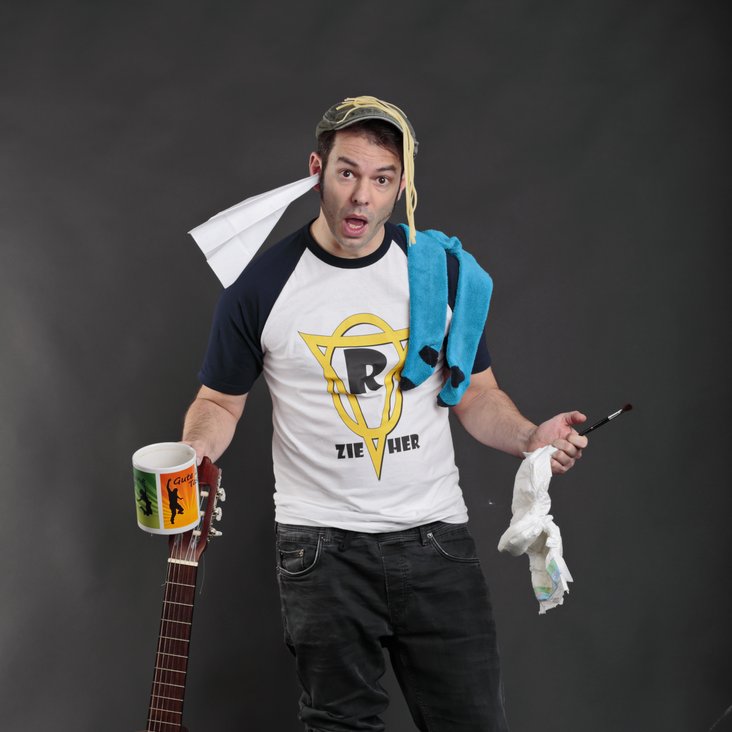 Yves Macak posiert mit einer Gitarre, einer Tasse, Windeln und einem Papierflieger im Ohr und schaut überfordert