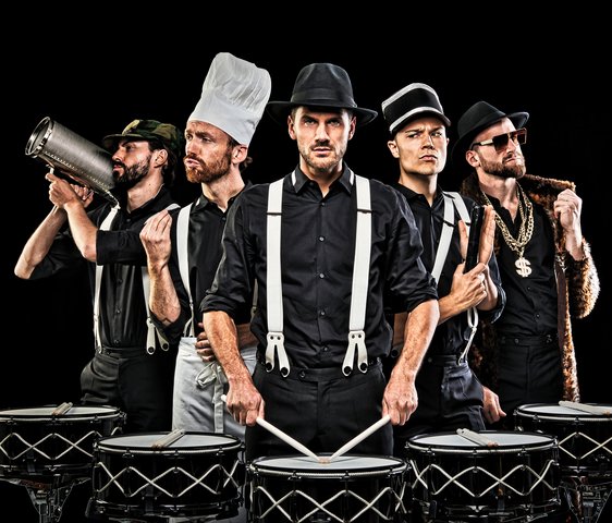 5 Männer stehen vor schwarzen Hintergrund vor Drums. Jeder von ihnen trägt eine andere Uniform
