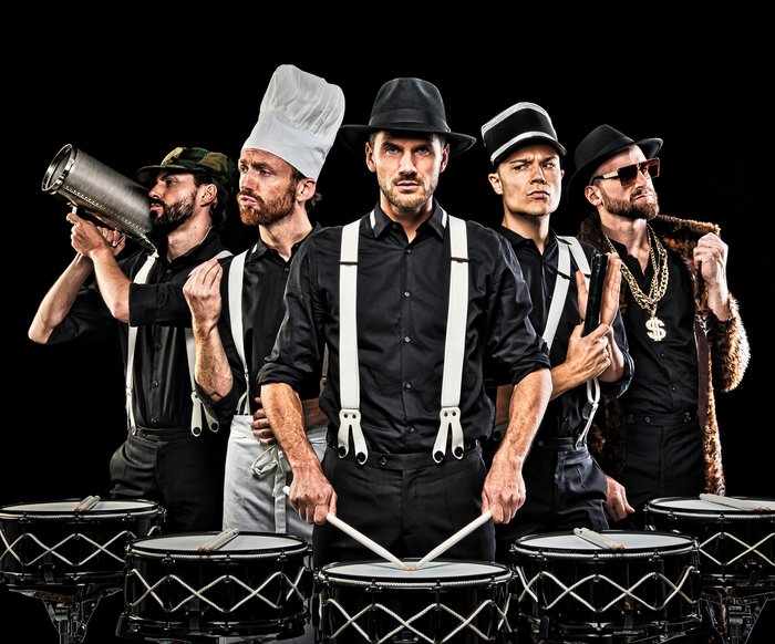5 Männer stehen vor schwarzen Hintergrund vor Drums. Jeder von ihnen trägt eine andere Uniform