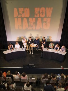 Publikum vor Bühne mit Podiumsrunde, auf Leinwand steht „AND NOW HANAU“.