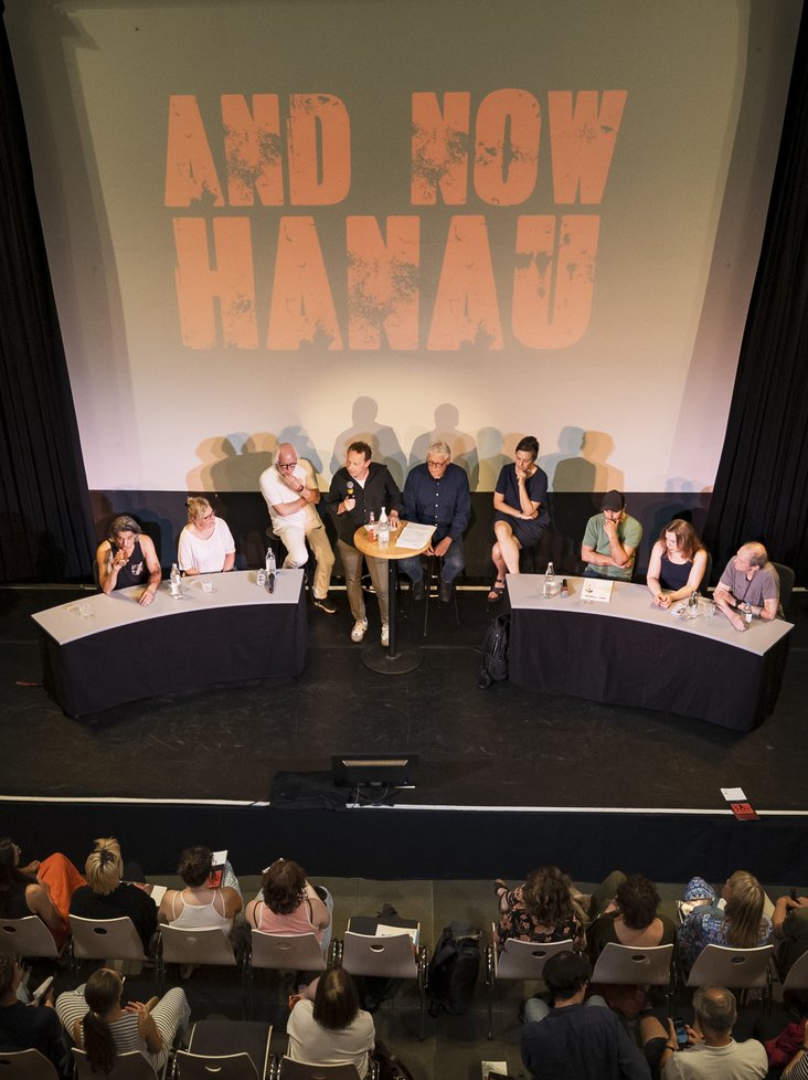 Publikum vor Bühne mit Podiumsrunde, auf Leinwand steht „AND NOW HANAU“.
