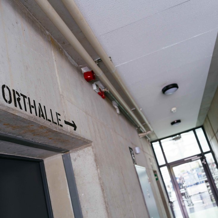 Flur mit Wegweiser „SPORTHALLE →“ an Betonwand.