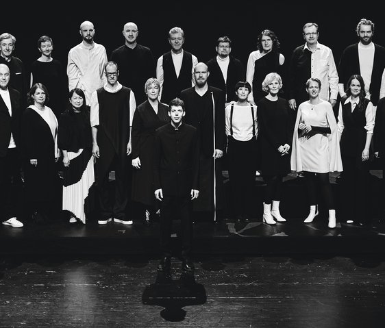 Das Vokalensemble steht positioniert für ein Gruppenbild auf einem Holzboden. Ein Mann steht vor den Reihen der anderen. Das Bild ist schwarz-weiß.