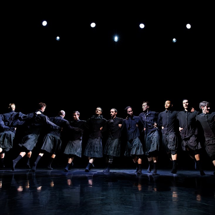 Die Tänzer:innen von Gauthier Dance in schwarzen Kostümen auf der Bühne. Die Bühne ist auch dunkel und es gibt wenig Licht.