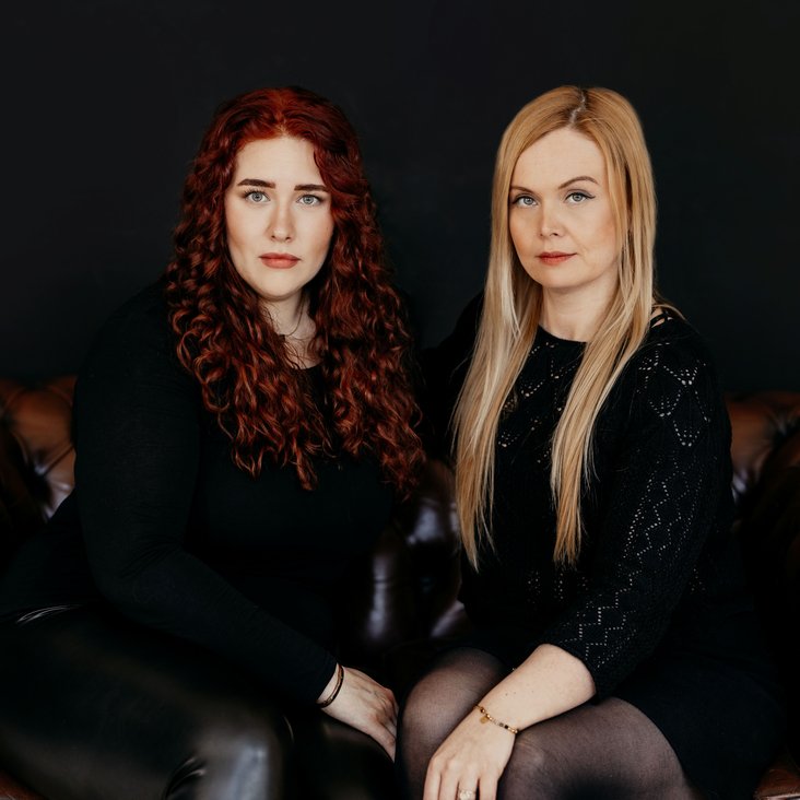 Portraitfoto der beiden Macherinnen von "Stimmen im Kopf". Sie sitzen auf einem braunen Ledersofa, tragen schwarze Kleidung und blicken in die Kamera.