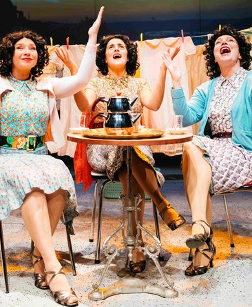 Drei Frauen sitzen an einem kleinen Tisch, alle heben beiden Hände in die Höhe. Sie scheinen zu singen oder ähnliches. Vor ihnen steht Tee, Wäsche im Hintergrund