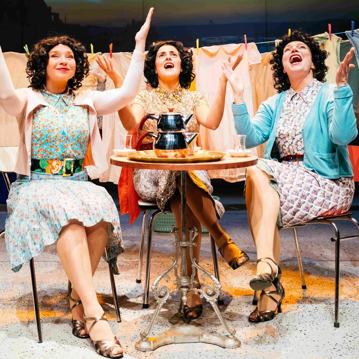 Drei Frauen sitzen an einem kleinen Tisch, alle heben beiden Hände in die Höhe. Sie scheinen zu singen oder ähnliches. Vor ihnen steht Tee, Wäsche im Hintergrund