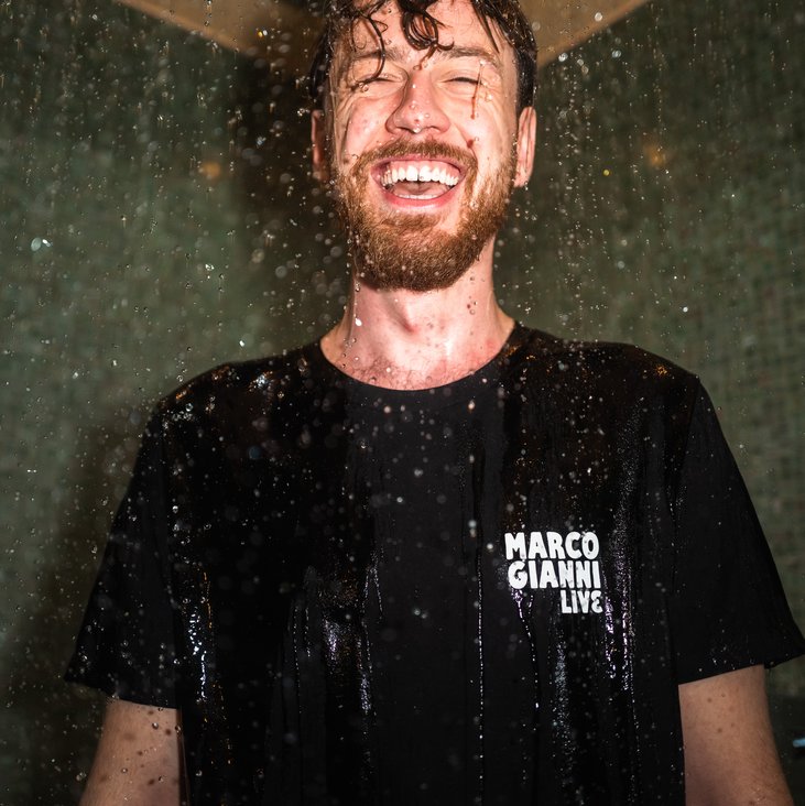 Marco Gianni steht unter einer Dusche in Klamotten und lacht