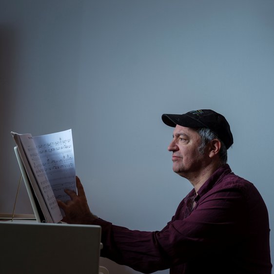 Rainald Grebe sitzt am Klavier, er blättert in Noten. Über ihm eine Deckenlampe die warmes Licht auf ihn wirft.