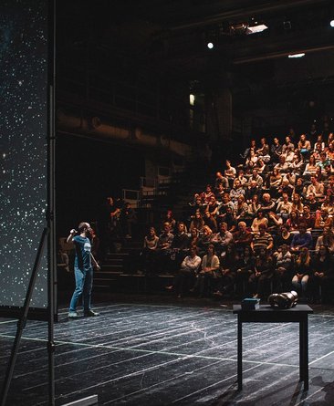 Das Foto zeigt die Veranstaltung Science Slam: Verschiedene Menschen sind auf der Bühne zu sehen, eine:r scheint etwas an einer Leinwand zu erklären, auf der das Universum oder ähnliches zu sehen ist. Im Hintergrund ist Publikum zu sehen.