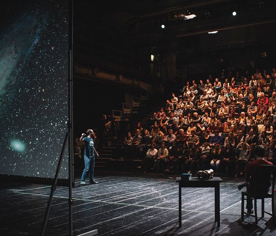Das Foto zeigt die Veranstaltung Science Slam: Verschiedene Menschen sind auf der Bühne zu sehen, eine:r scheint etwas an einer Leinwand zu erklären, auf der das Universum oder ähnliches zu sehen ist. Im Hintergrund ist Publikum zu sehen.