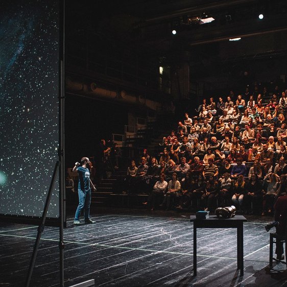 Das Foto zeigt die Veranstaltung Science Slam: Verschiedene Menschen sind auf der Bühne zu sehen, eine:r scheint etwas an einer Leinwand zu erklären, auf der das Universum oder ähnliches zu sehen ist. Im Hintergrund ist Publikum zu sehen.