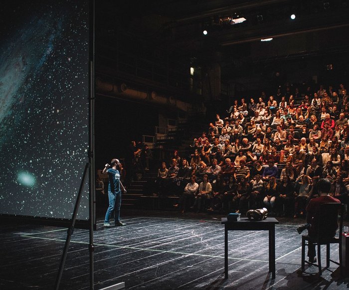 Das Foto zeigt die Veranstaltung Science Slam: Verschiedene Menschen sind auf der Bühne zu sehen, eine:r scheint etwas an einer Leinwand zu erklären, auf der das Universum oder ähnliches zu sehen ist. Im Hintergrund ist Publikum zu sehen.