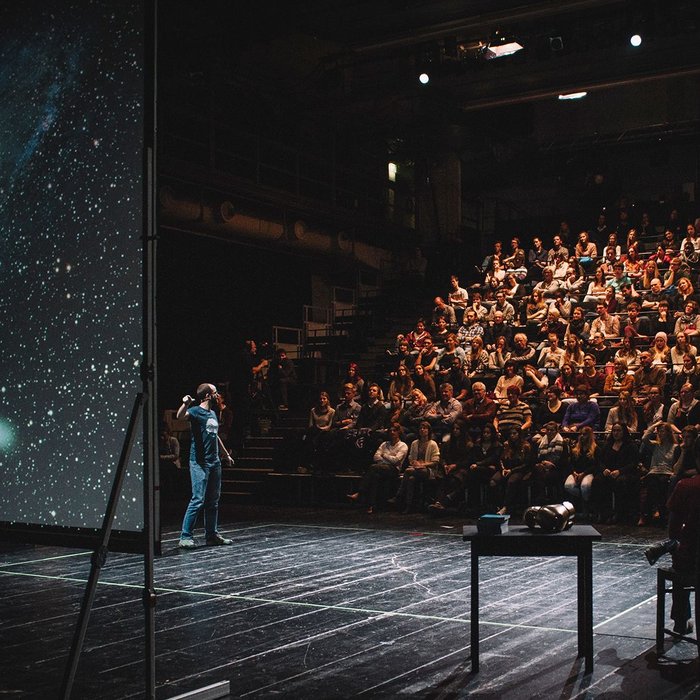 Das Foto zeigt die Veranstaltung Science Slam: Verschiedene Menschen sind auf der Bühne zu sehen, eine:r scheint etwas an einer Leinwand zu erklären, auf der das Universum oder ähnliches zu sehen ist. Im Hintergrund ist Publikum zu sehen.