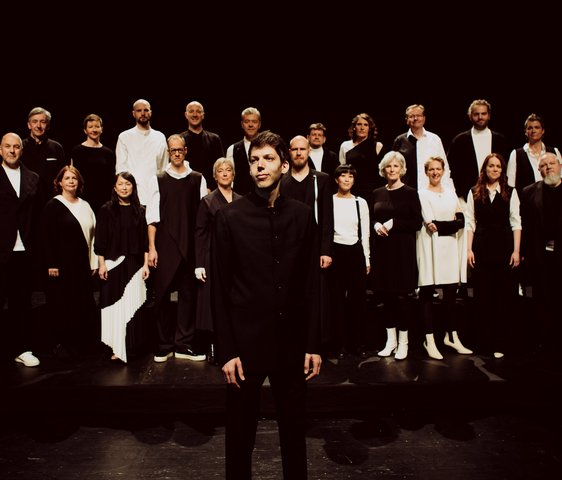 Großes Vokalensemble auf einer Bühne, Gruppenaufstellung unter Bühnenlicht.