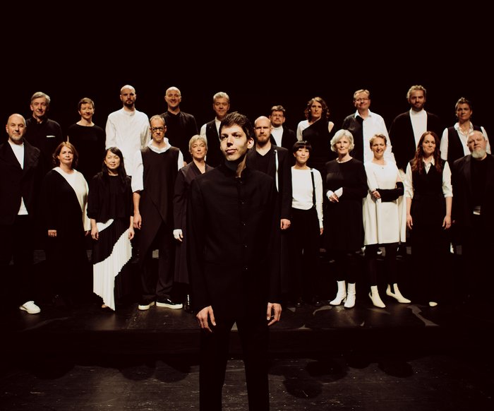Großes Vokalensemble auf einer Bühne, Gruppenaufstellung unter Bühnenlicht.