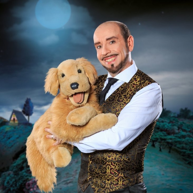 Der Künstler hat einen Stoffhund auf dem Arm und grinst in die Kamera.