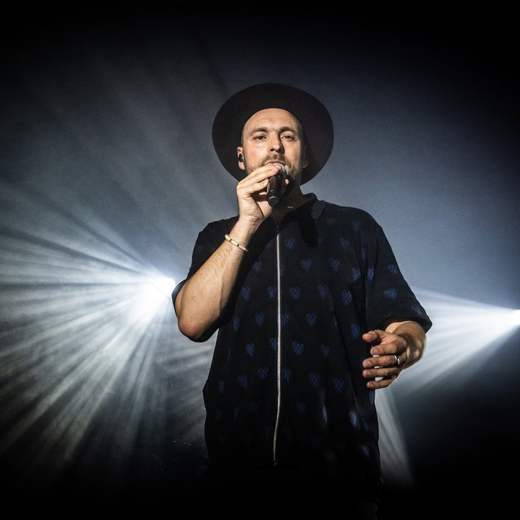 Max Mutzke trägt einen Hut und singt in ein Mikrofon, im Hintergrund ist das Licht eines Scheinwerfers zu sehen.