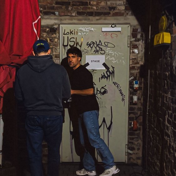 Moritz Hohl vor einer Stahltür mit vielen Tags, das Setting sieht aus wie ein Veranstaltungsort oder Backstagebereich.