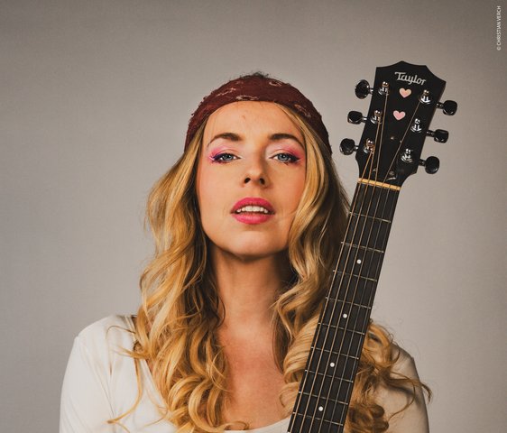 Eine Frau posiert mit einer Gitarre. Sie hat blonde Haare und ein rotes Tuch um den Kopf gewickelt.