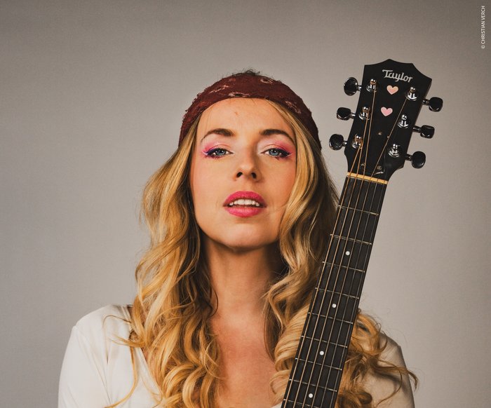 Eine Frau posiert mit einer Gitarre. Sie hat blonde Haare und ein rotes Tuch um den Kopf gewickelt.