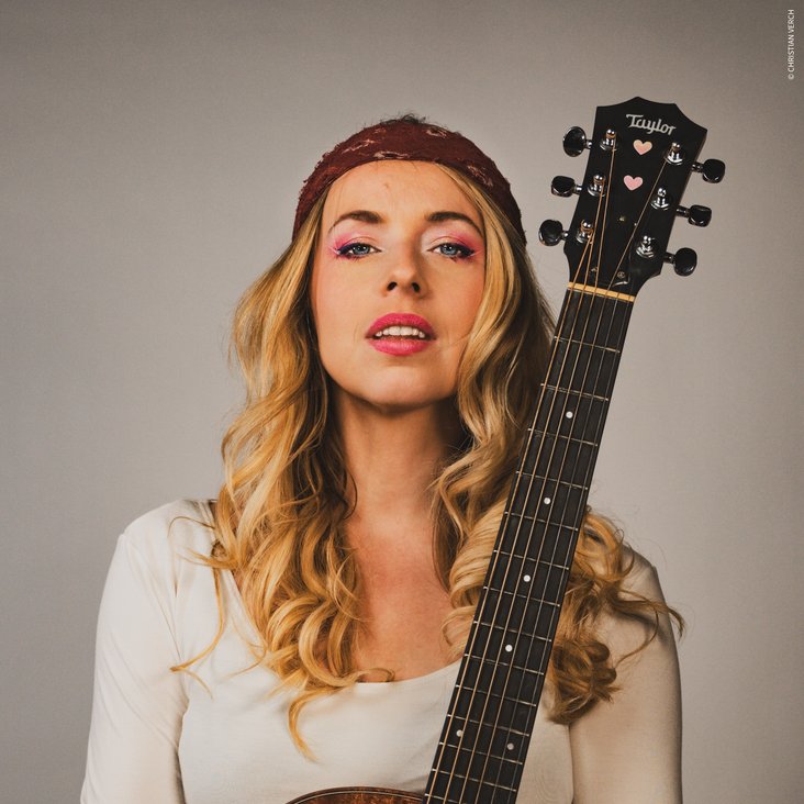 Eine Frau posiert mit einer Gitarre. Sie hat blonde Haare und ein rotes Tuch um den Kopf gewickelt.