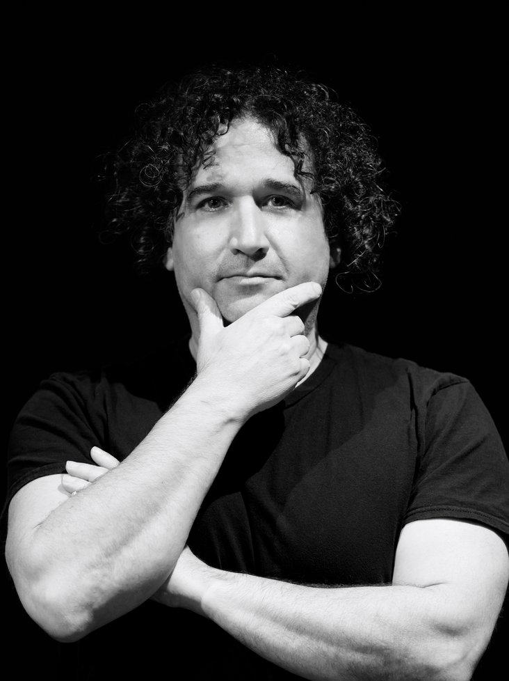 Ein Portrait des Choreographen Barak Marshall in schwarz-weiß. Er trägt ein schwarzes Shirt und hat dunkle, lockige Haare.