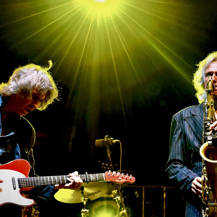 Die beiden Musiker der Band "The Dire Straits Experience" stehen gemeinsam auf der Bühne. Auf der linken Seiten spielt der Musiker auf einer weiß-roten E-Gitarre und ist am headbangen. Der Musiker auf der rechten Seite spielt Saxophon. Beide Musiker haben schulterlanges Haar und tragen blaue Jacken. Der Hintergrund ist schwarz und am oberen Bildrand in der Mitte strahlt ein gelb-grüner Scheinwerfer.