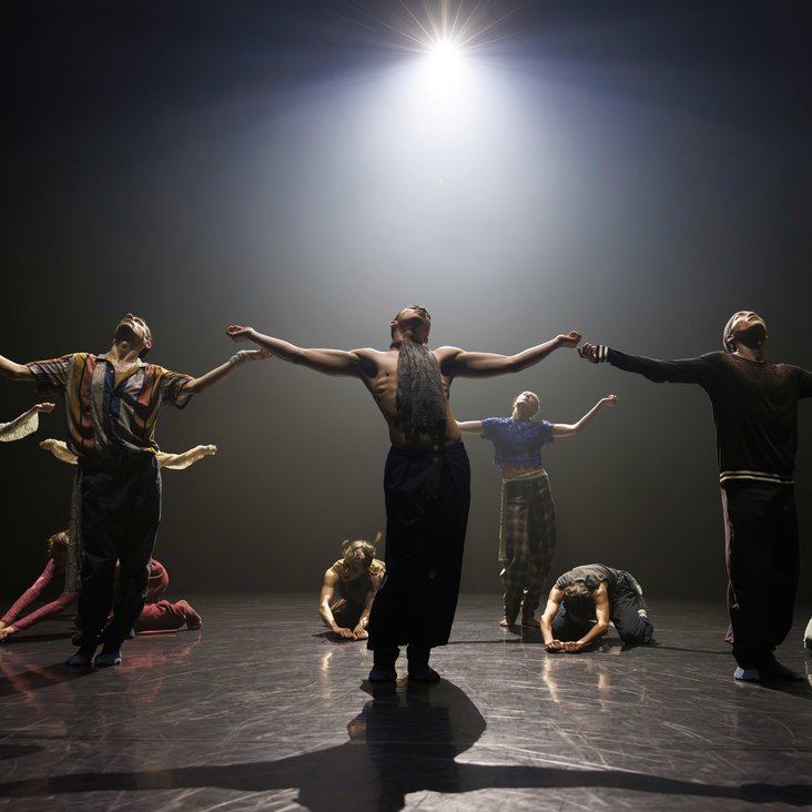 Die Tänzer:innen von Gauthier Dance in verschiedenfarbigen Kostümen auf der Bühne. Die Bühne ist auch dunkel und die Beleuchtung ist atmosphärisch.