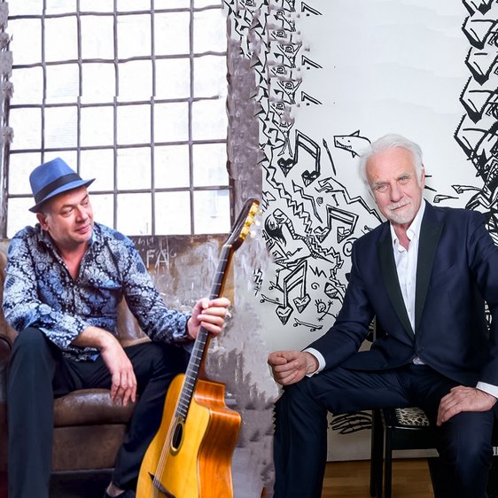 Zwei Musiker sitzen einander gegenüber, getrennt durch eine vertikale Bildkante; zwischen ihnen steht eine Akustikgitarre.