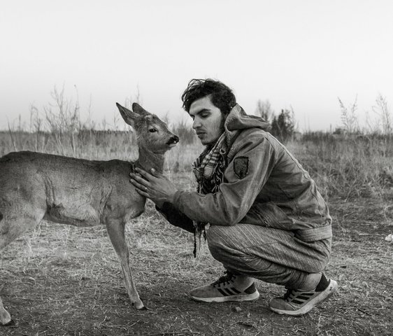 Ein schwarz-weiß Foto von einem Mann, der in der Hocke sitzt. Er hat die Augen geschlossen. Vor ihm steht ihm zugewandt ein Hirschweibchen, das er am Hals streichelt.
