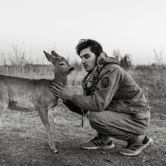 Ein schwarz-weiß Foto von einem Mann, der in der Hocke sitzt. Er hat die Augen geschlossen. Vor ihm steht ihm zugewandt ein Hirschweibchen, das er am Hals streichelt.