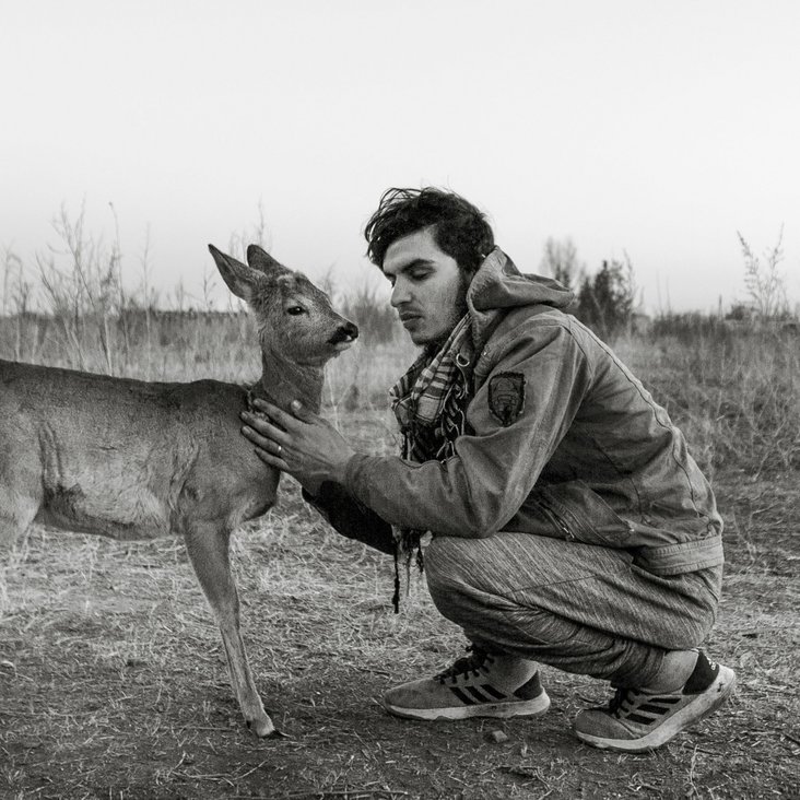 Ein schwarz-weiß Foto von einem Mann, der in der Hocke sitzt. Er hat die Augen geschlossen. Vor ihm steht ihm zugewandt ein Hirschweibchen, das er am Hals streichelt.