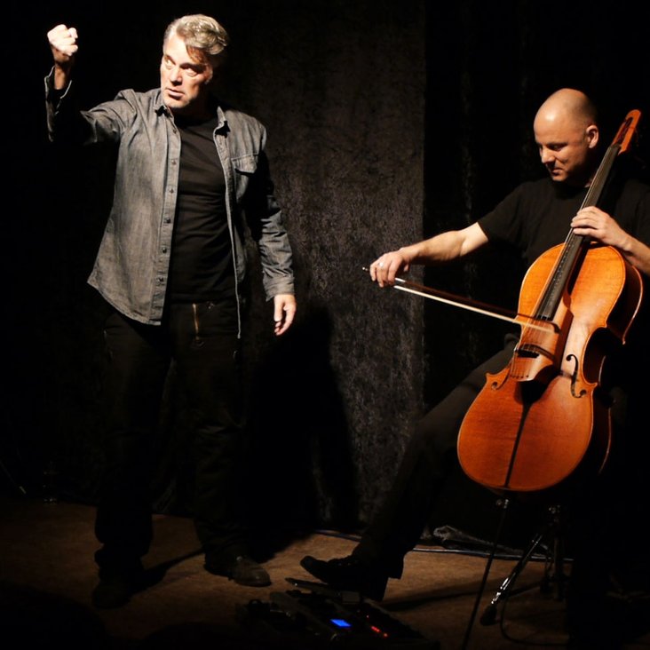 Zwei Männerauf der Bühne. Der eine schauspielert, der andere spielt Cello.