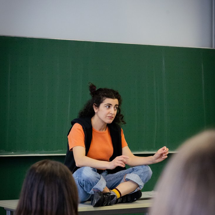 hauspielerin sitzt in einem Klassenzimmer auf einem Tisch vor einer Schultafel. Man sieht die Schüler:innen, die dem Klassenzimmerstück zusehen, von hinten.