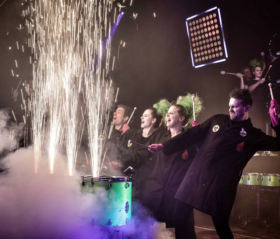 Das greenbeats Ensemble auf der Bühne, sie trommeln auf Tonnen, aus denen feuerwerksähnliche Funken sprühen.