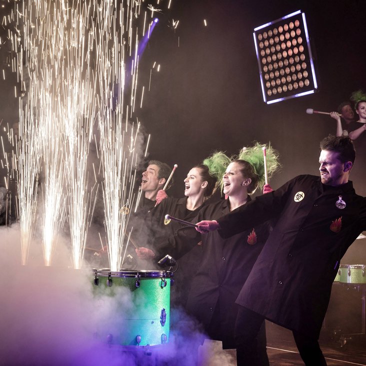Das greenbeats Ensemble auf der Bühne, sie trommeln auf Tonnen, aus denen feuerwerksähnliche Funken sprühen.