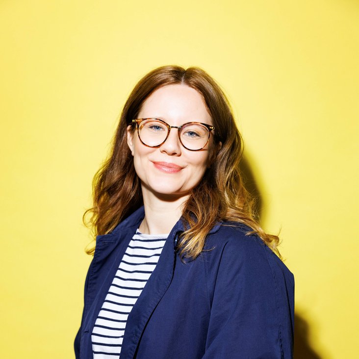 Leonie Schöler trägt ein blau-weiß gestreiftes Oberteil und darüber einen dunkelblauen Blazer. Sie trägt eine Brille und dreht ihr Gesicht lächelnd in die Kamera. Sie steht vor einer gelben Wand.
