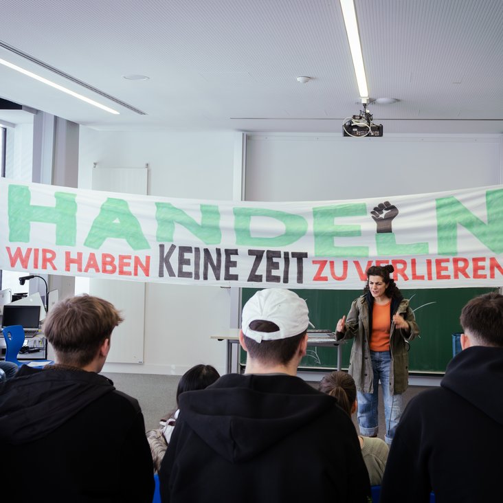 Ein Schauspielerin steht in einem Klassenzimmer vor einer Tafel. Hinter ihr stehen zwei Schüler:innen, die ein Transparent halten. Man sieht die Schüler:innen, die dem Klassenzimmerstück zusehen, von hinten.