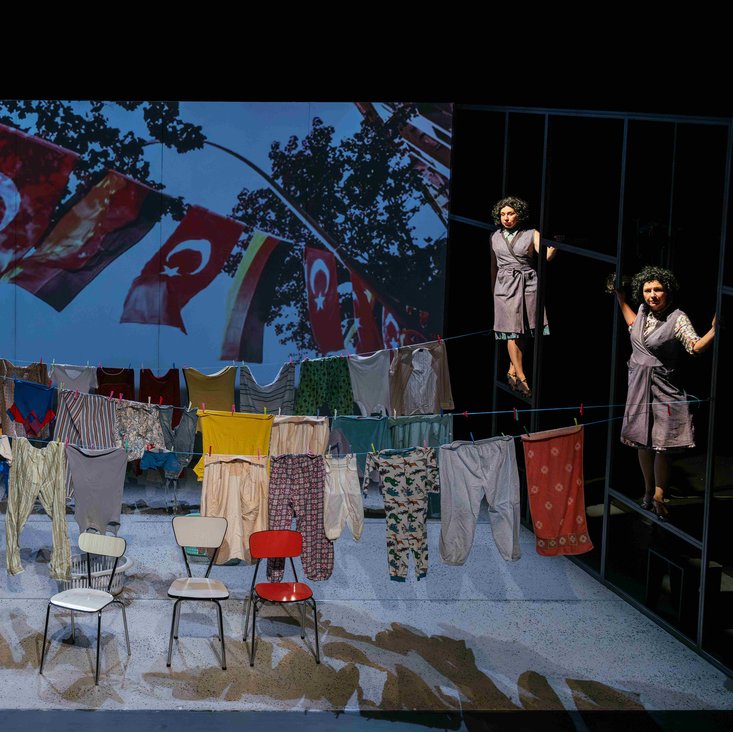 Bühnenszene mit mehreren Schauspielerinnen und Wäscheleinen, im Hintergrund die Projektion eines Fotos auf dem türkische Flaggen zu sehen sind