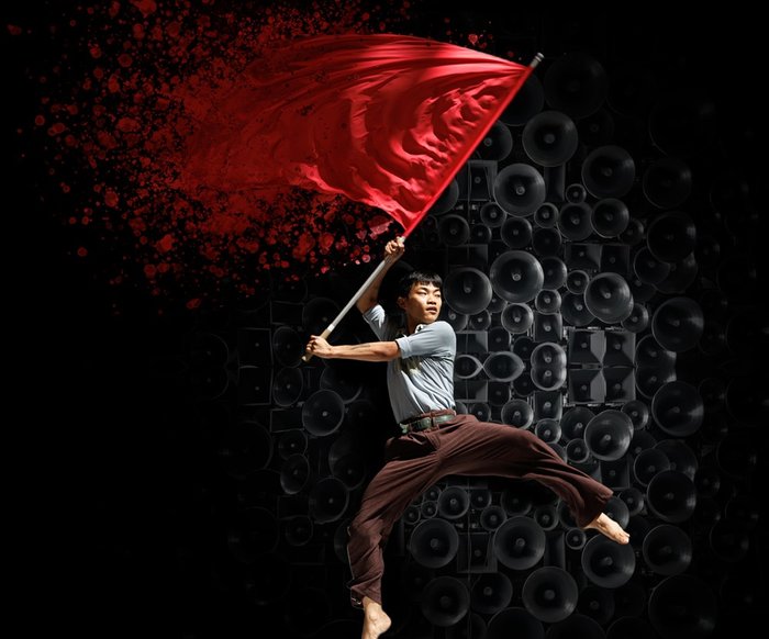 Ein Tänzer schwingt im Sprung eine rote Flagge, welche sich am Ende auflöst. Im Hintergrund sind Lautsprecher.