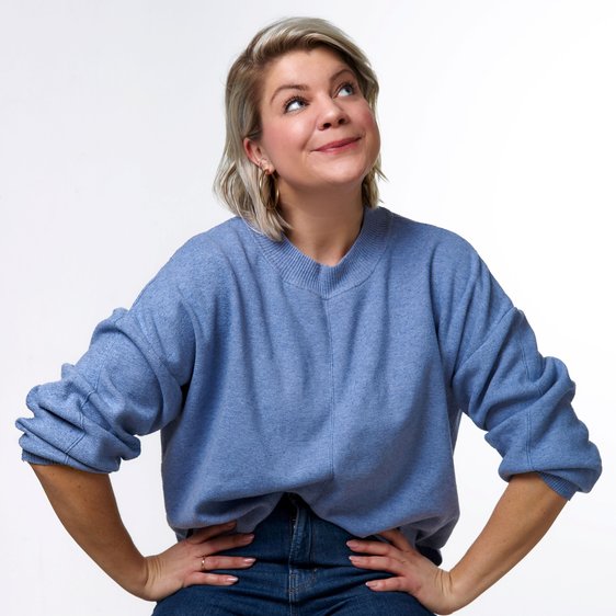Portraitfoto von Frieda Lewin. Sie sitzt auf einem Barhocker, trägt einen blauen Pullover und Jeans, hat die Hände in die Hüften gelegt und schaut nach oben.