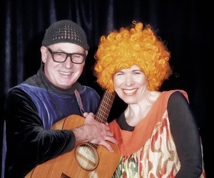 Christof und Vladi Altmann auf der Bühne. Er trägt ein langes lilafarbenes Kostüm und hat eine Gitarre in der Hand. Sie trägt ein buntes Kleid und eine Perücke mit kurzen orangefarbenen Locken.