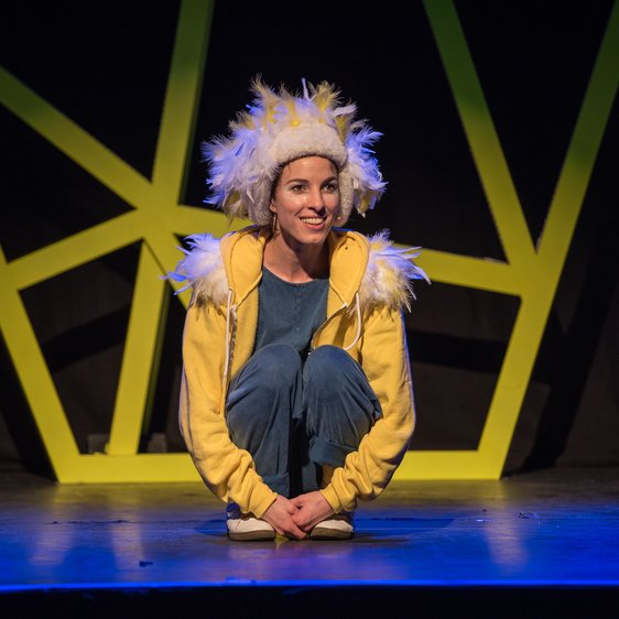 Bühnenfoto "Eule findet den Beat". Eine Sängerin in gelb-blauer Kleidung und Eulen-Feder-Kostüm auf einer Theaterbühne.