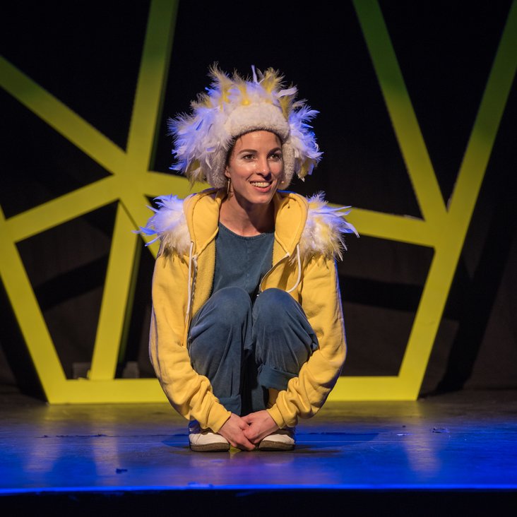 Bühnenfoto "Eule findet den Beat". Eine Sängerin in gelb-blauer Kleidung und Eulen-Feder-Kostüm auf einer Theaterbühne.