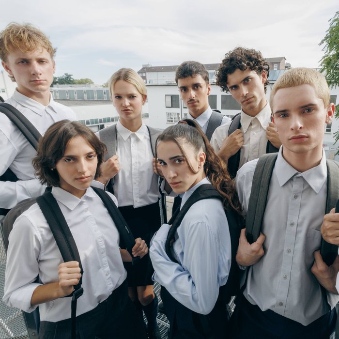 Die Tänzer:innen der Gauthier Dance JUNIORS stehen eng beieinander in hellen Hemden und Blusen mit Rucksäcken und blicken direkt in die Kamera.