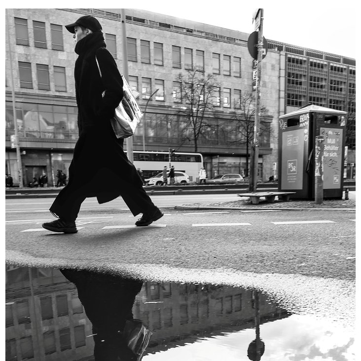 Ein Mensch geht durch eine Straße von Berlin und spiegelt sich dabei in einer Pfütze.