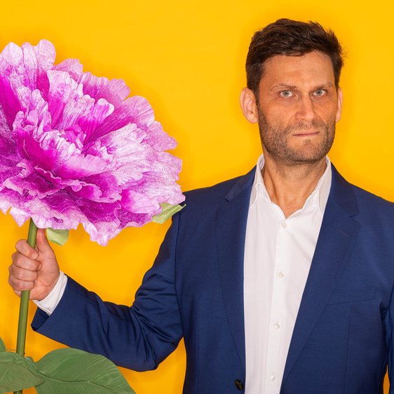 Portraitfoto Christian Ehring. Er trägt einen blauen Anzug und hält eine überdimensional große Blume in der Hand.