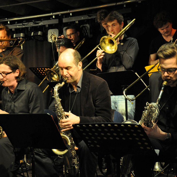 Die Big Band mit Instrumenten auf der Bühne, die Musiker:innen tragen schwarze Kleidung