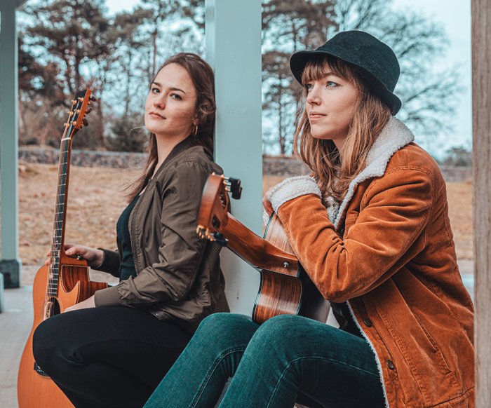 Portaitfoto Sophie Chassée und Karlijn Langendijk. Sie sitzen draußen unter einem Vordach und haben beide Gitarren in der Hand.